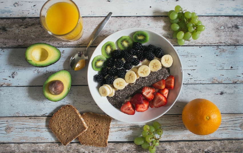 Gesunde Gewohnheiten im Alltag: Chia-Bowl mit Banane Beeren und Kiwi dazu Avocado Vollkornbrot Trauben und ein Glas Orangensaft auf Holztisch